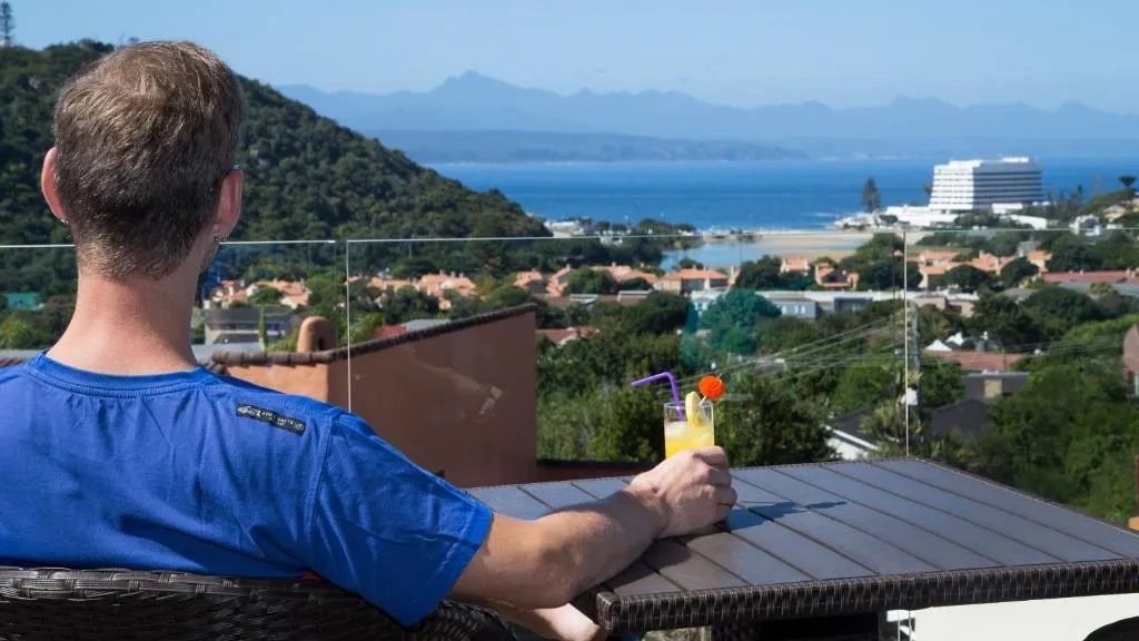 Guest relaxing on a balcony with a drink, overlooking Plettenberg Bay's beach and mountains