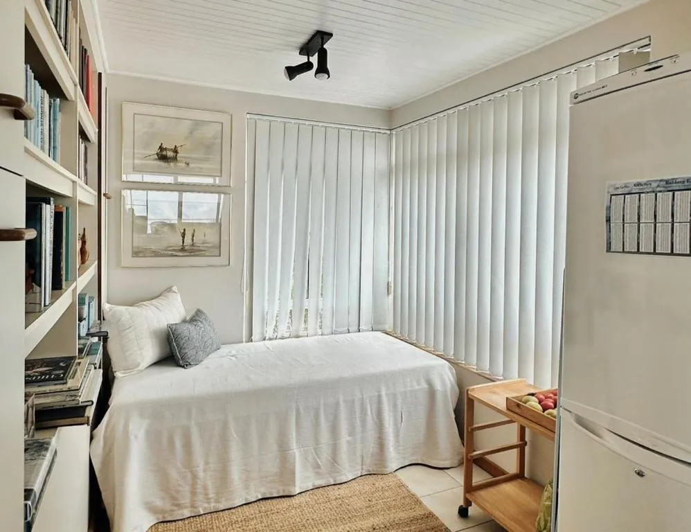 Single bedroom with built-in bookshelves and vertical blinds beside a compact fridge