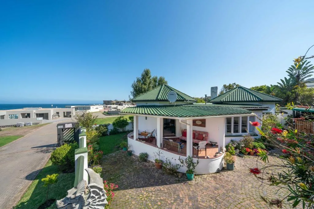 White cottage with green pyramid roofs, a curved veranda, garden, and the ocean visible beyond