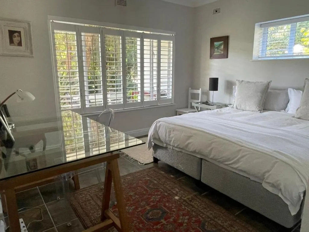 Bedroom with a glass desk, upholstered bed and shuttered windows overlooking greenery