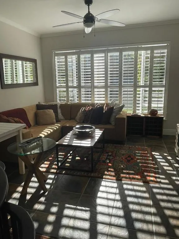Living room with plantation shutters, a ceiling fan and a patterned rug on dark tile flooring