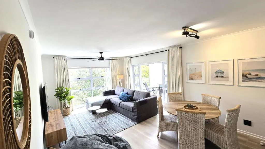 Open-plan lounge with grey corner sofa, round dining table and full-height windows onto greenery