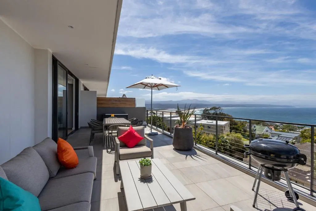 Balcony lounge with orange and red cushions, a gas braai and umbrella-shaded dining table over the sea