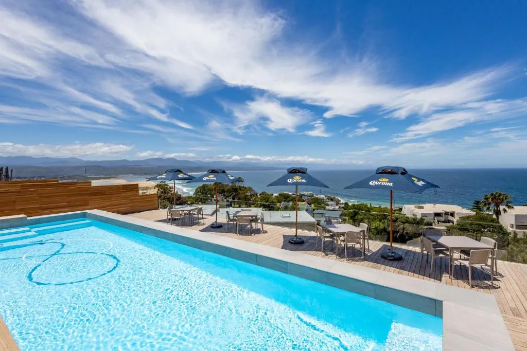 Rooftop infinity pool with a wooden deck and umbrella-shaded loungers overlooking the ocean and coastline