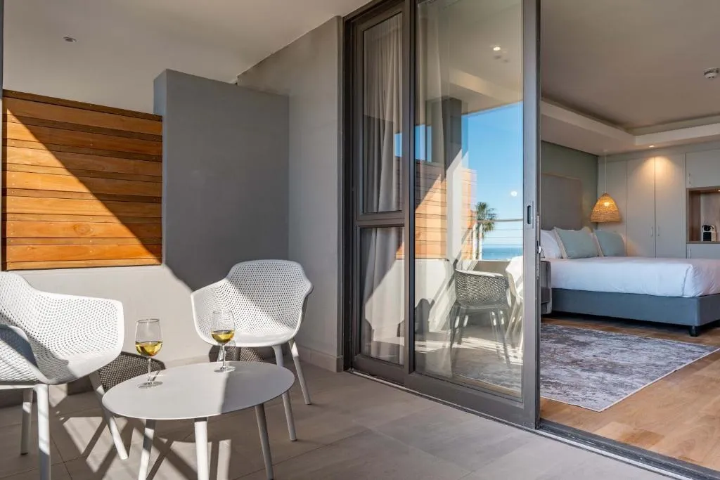 Hotel room with sliding doors onto a balcony seating area, the sea visible beyond the bed