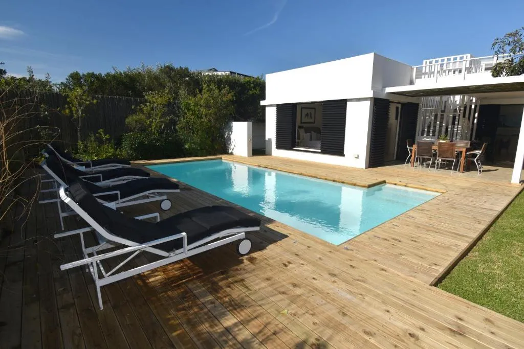 Wooden pool deck with black sun loungers facing the pool, house and covered outdoor dining area
