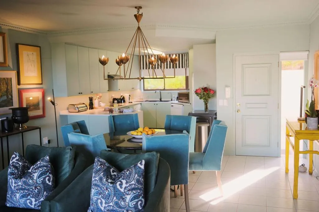 Round dining table with teal velvet chairs beneath a brass flower-form chandelier, open to the kitchen