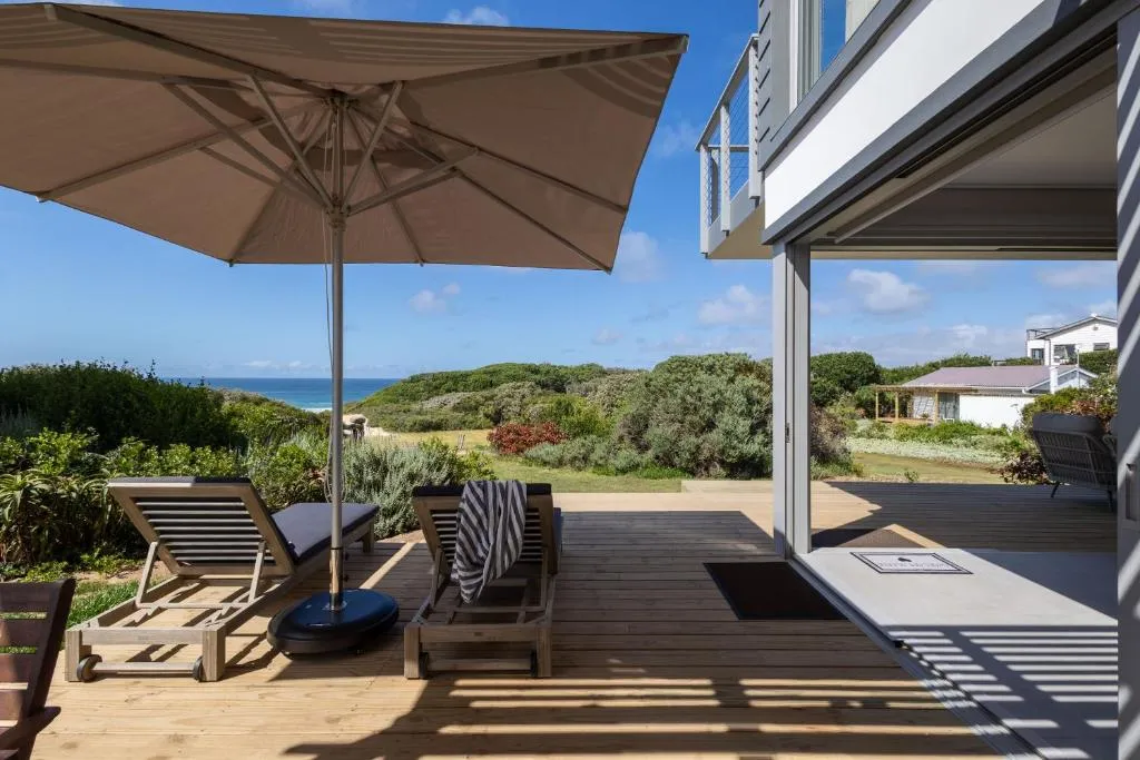 Wooden deck with a shade umbrella and loungers overlooking dune bush to a strip of sea