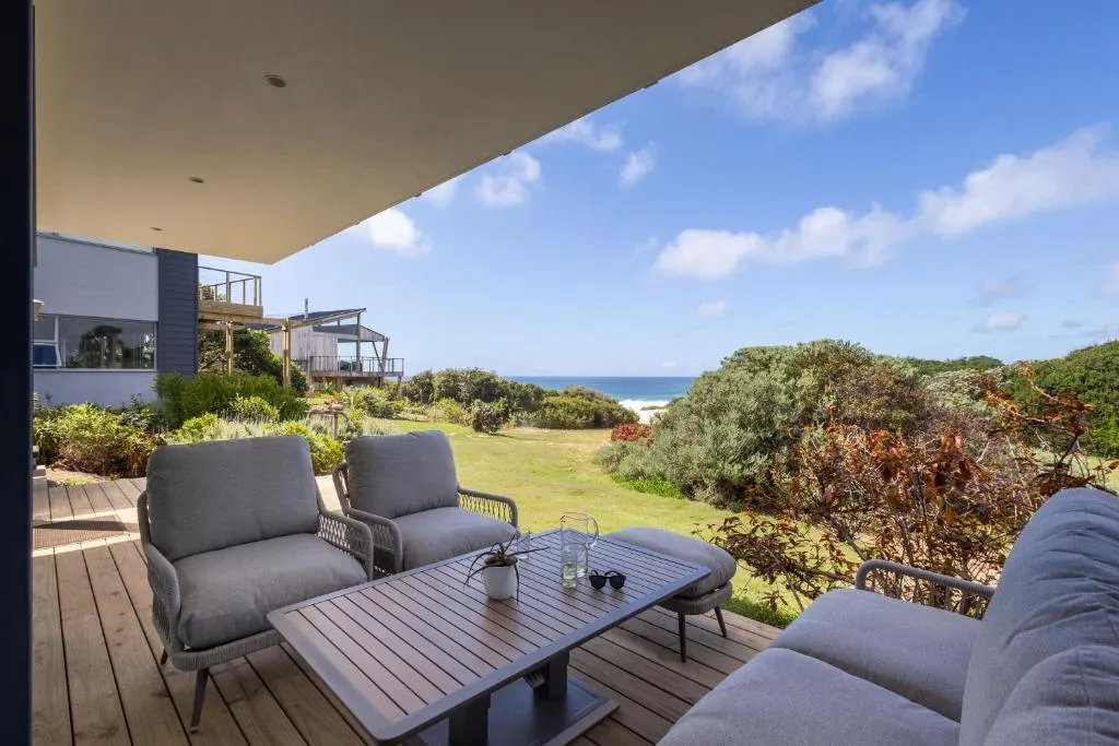 Covered patio seating with wicker armchairs looking out over dune bush to the beach and sea