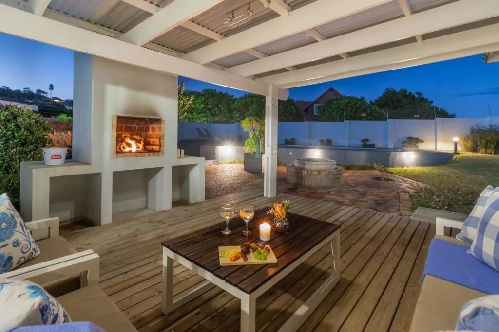 Covered wooden deck with a built-in braai and fire pit, wine glasses set beside a floodlit pool at dusk