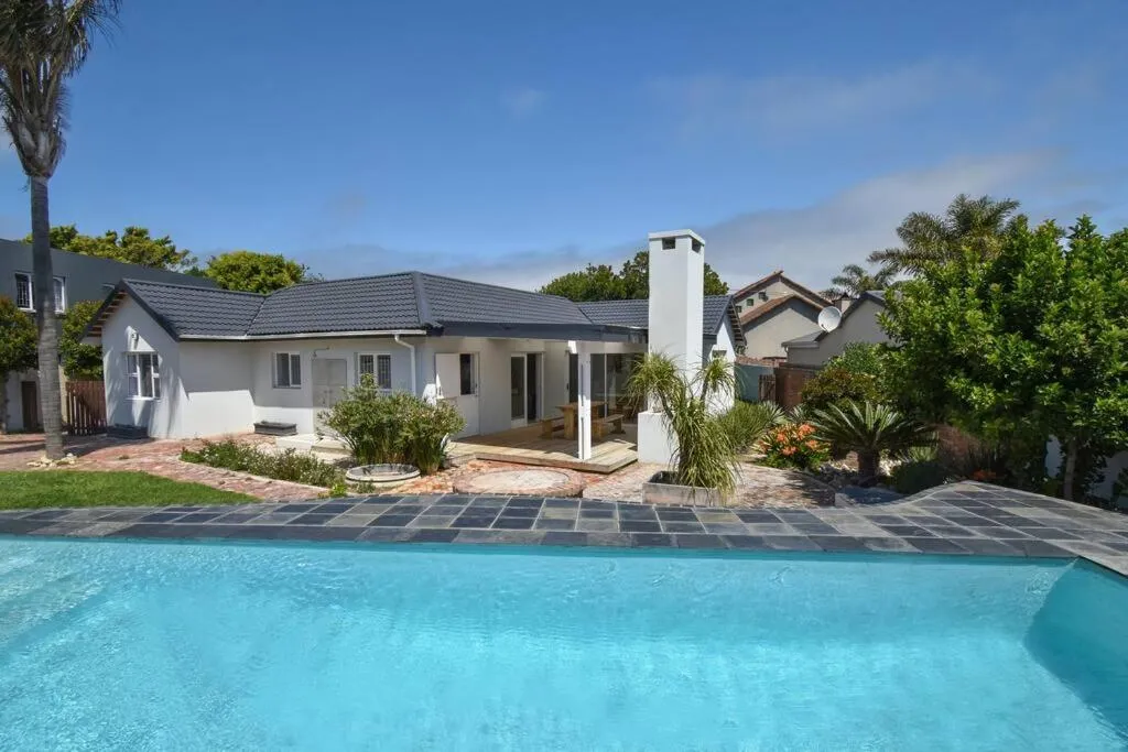 Single-storey house exterior with a long swimming pool, lawn and slate paving under a palm tree