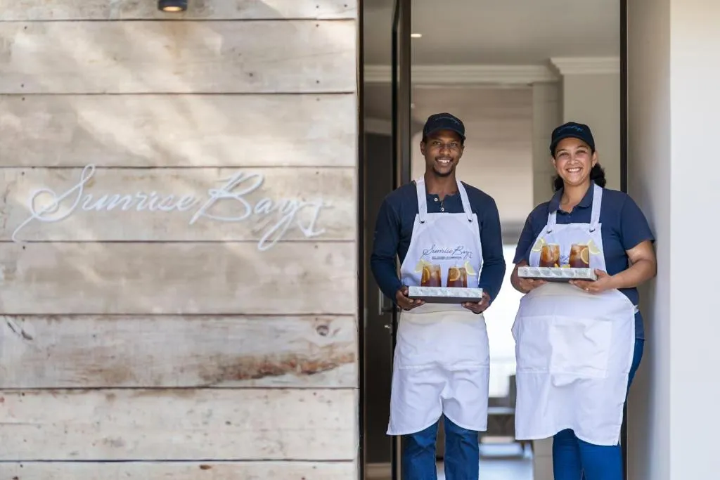Staff at the entrance holding welcome trays beside the carved Sunrise Bay timber sign