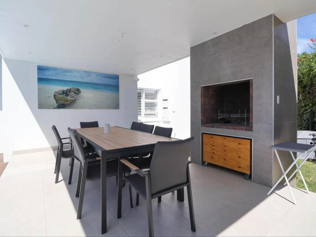 Covered outdoor dining table beside a built-in brick braai with a beach-themed painting
