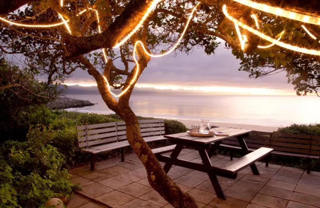 Wooden picnic table on a terrace strung with fairy lights, facing the sea at dusk