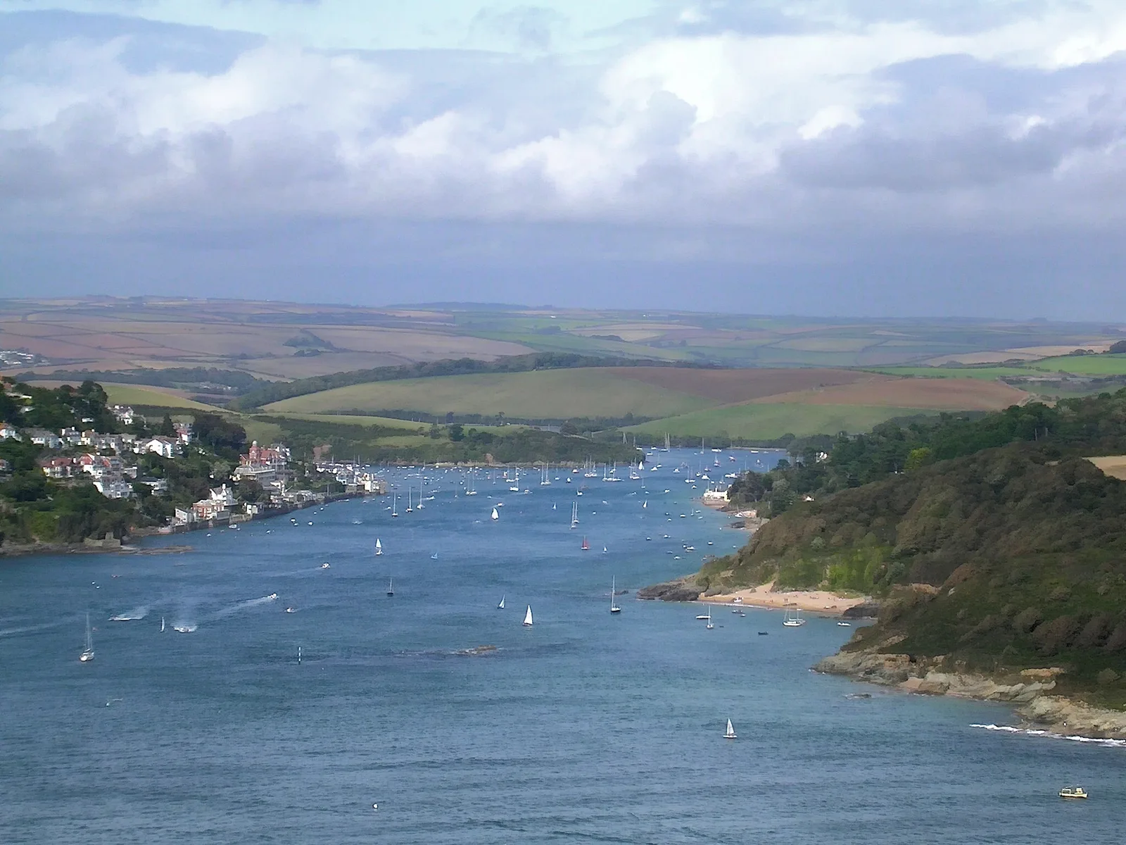 The Salcombe estuary seen from high ground at Sharpitor, looking inland — the town on the wooded left bank, dozens of moored yachts on the water, a sandy cove on the far shore at right, and rolling farmland ridges beyond