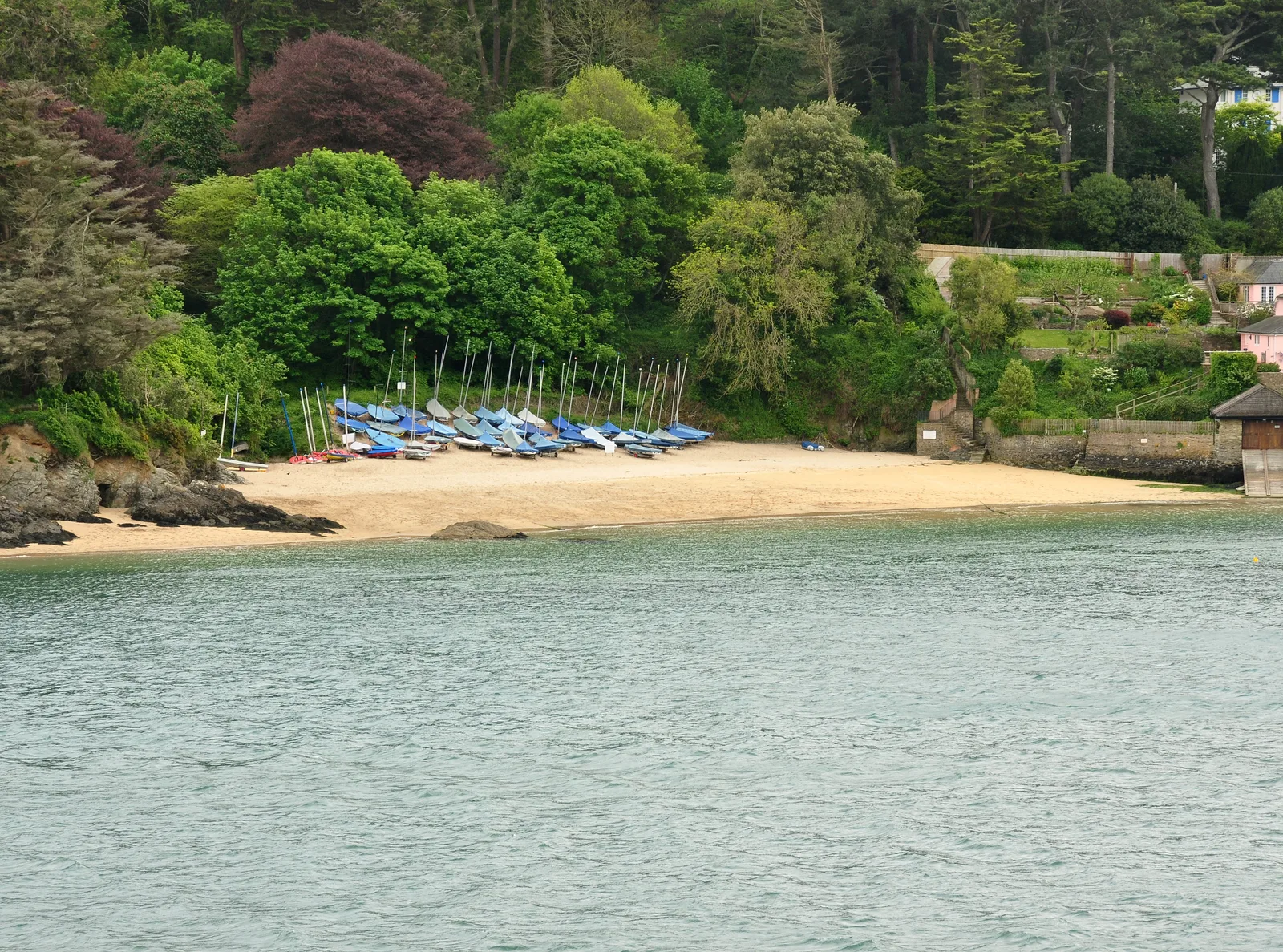 Mill Bay beach across the estuary from Salcombe, a small sandy cove with dinghies pulled up on the sand and trees behind