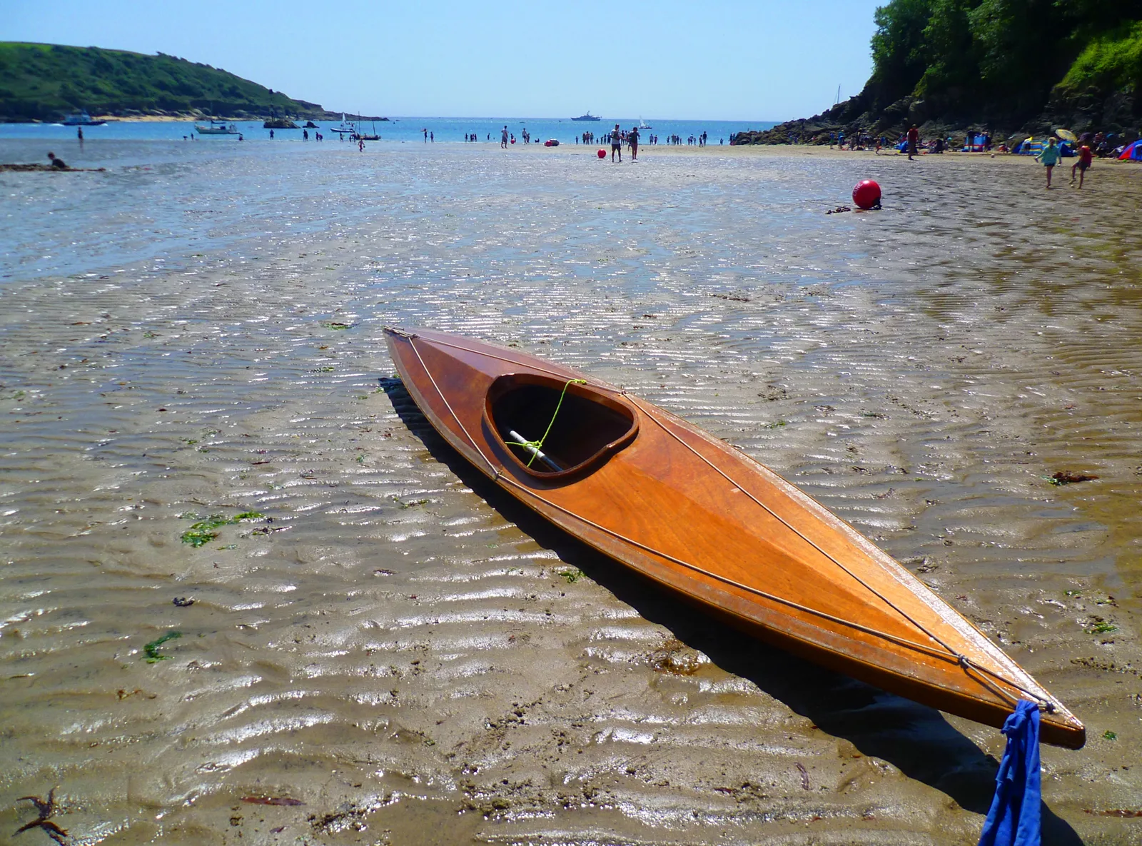 A varnished wooden kayak lying on rippled wet sand at low tide on North Sands, with people paddling in the shallows and boats moored off the beach in the distance