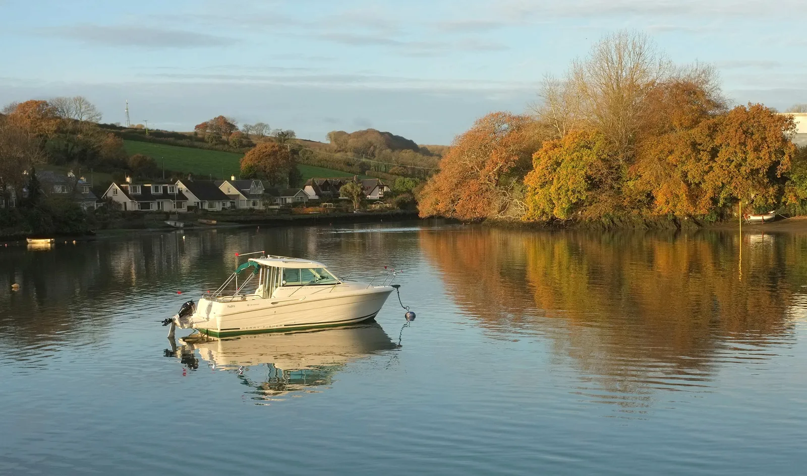A white motor cruiser moored on flat calm water at the Kingsbridge end of the estuary, with autumn trees in orange and gold on the bank and a row of houses on the hillside behind