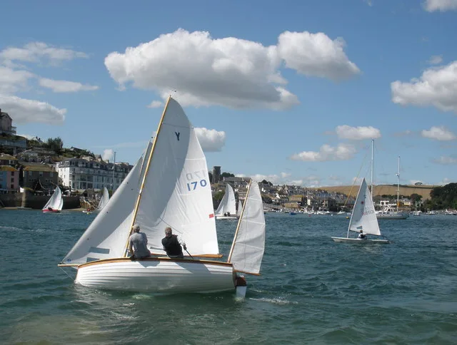 A white wooden sailing dinghy with two crew and cream sails racing across the harbour at Salcombe, with more small sailing boats behind it and the houses of the town on the hillside