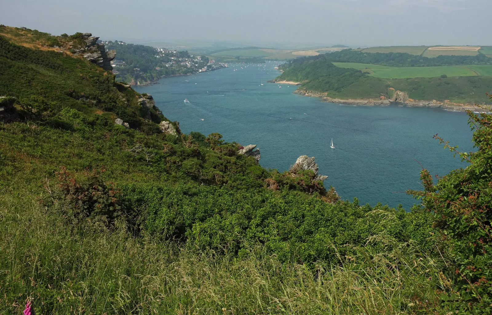 The mouth of the Salcombe estuary seen from high on the cliffs, with a white yacht under sail on deep blue water, wooded headlands on either side and the town of Salcombe in the distance at the head of the channel