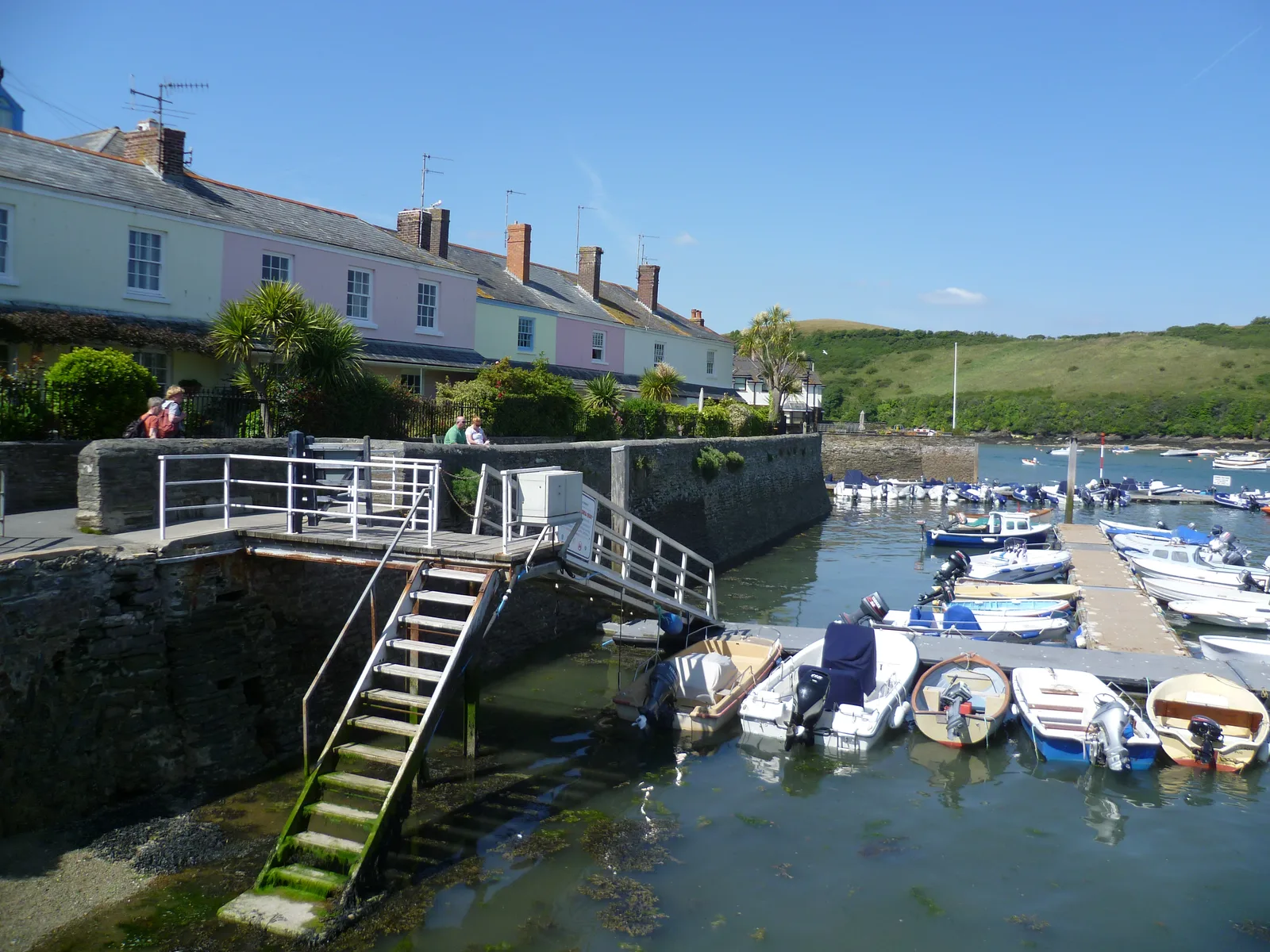 A row of small open boats with outboard motors moored alongside floating pontoons off a stone quay at Salcombe, with pastel-painted cottages behind and a green hillside across the water