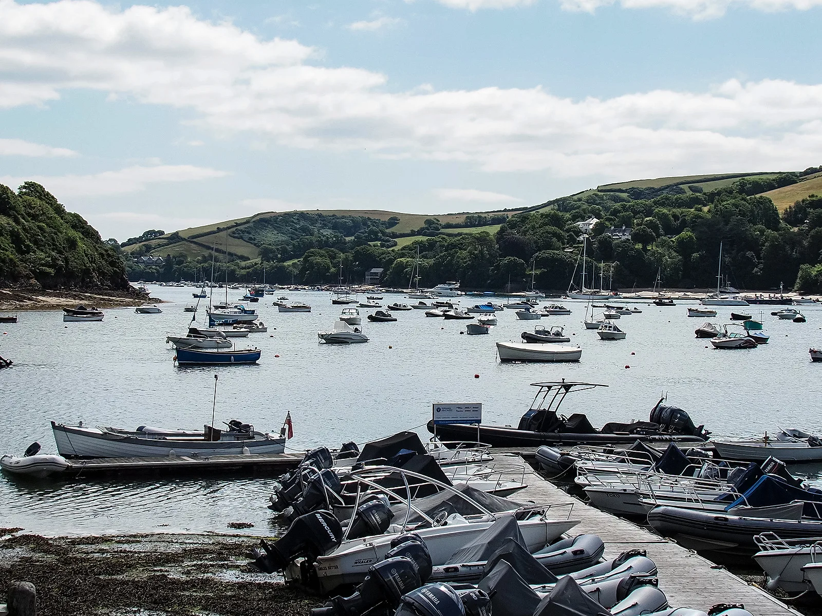 Moorings in Batson Creek at Salcombe, with dinghies and small motorboats drawn up on a pontoon in the foreground and moored boats on the water beyond, green hills rising behind