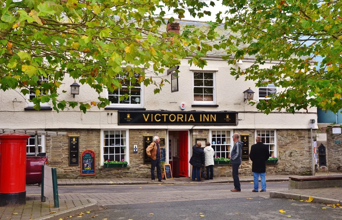 The frontage of the Victoria Inn in Salcombe, a cream and stone pub with a black sign reading Victoria Inn in gold lettering, sash windows, window boxes and people standing outside the red door