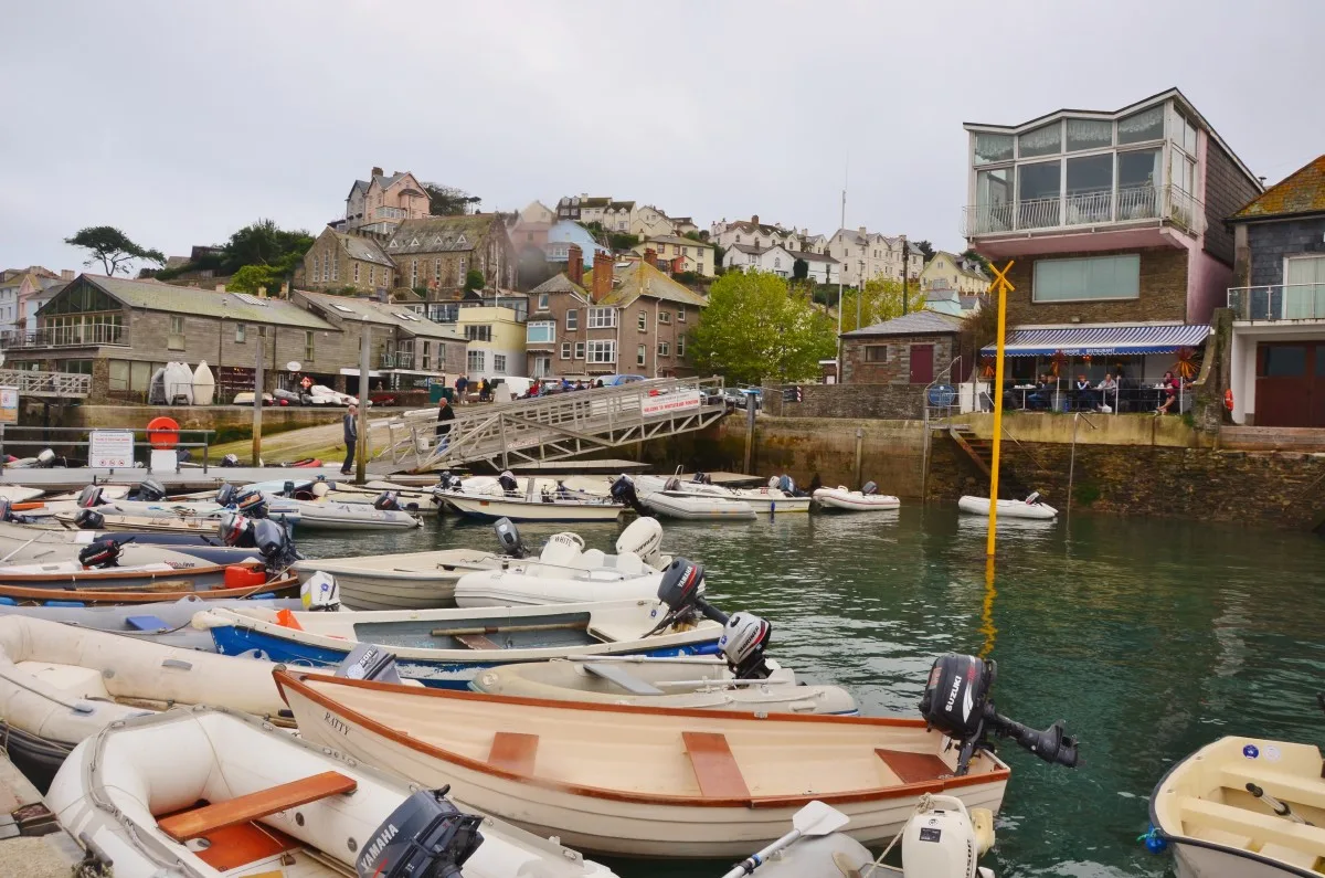 A waterside restaurant in Salcombe with a blue and white striped awning and people sitting at tables under it, dinghies moored on the water in front and the town rising on the hillside behind