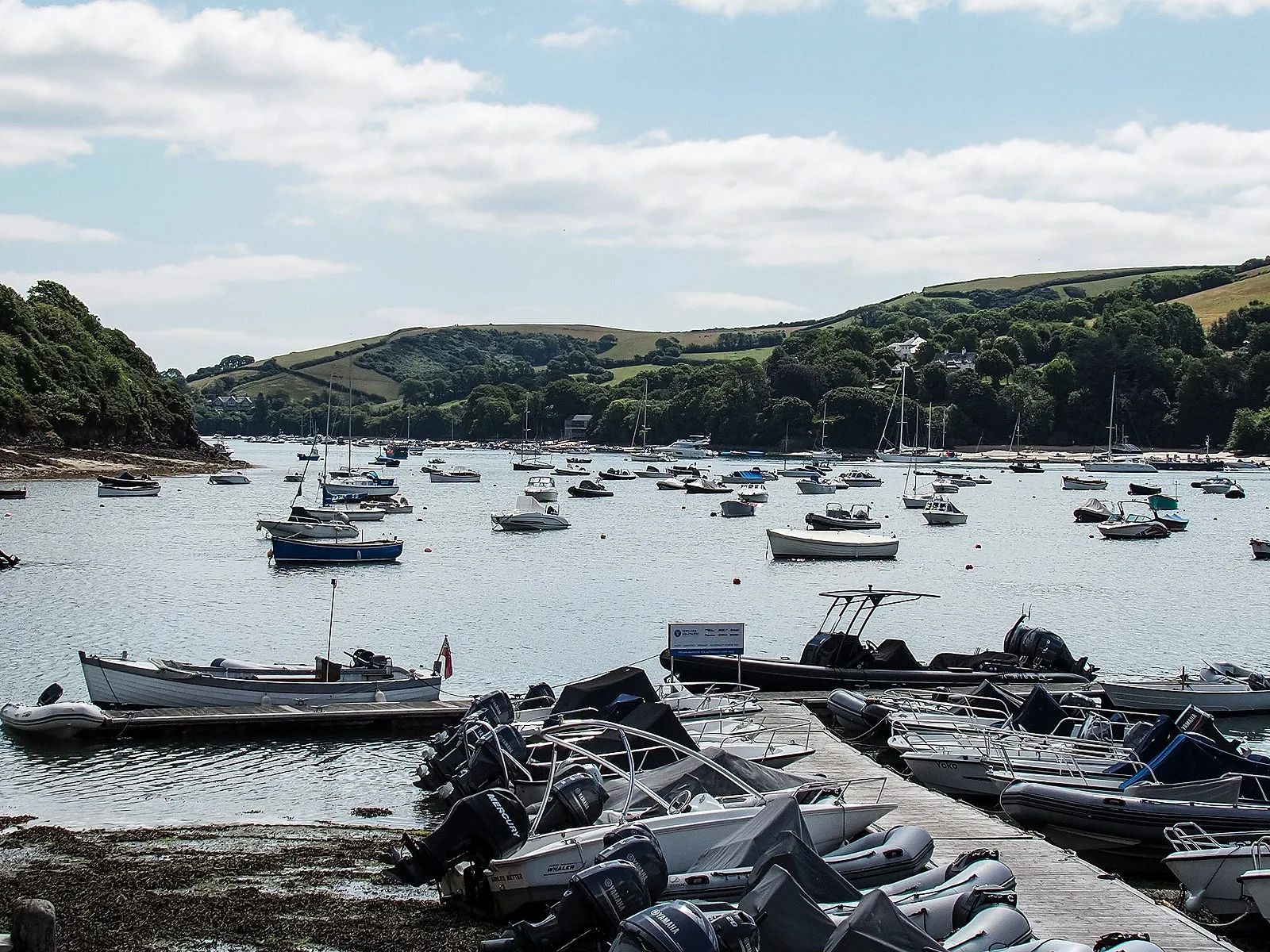Moorings in Batson Creek at Salcombe, with dinghies and small motorboats drawn up on a pontoon in the foreground and moored boats on the water beyond, green hills rising behind