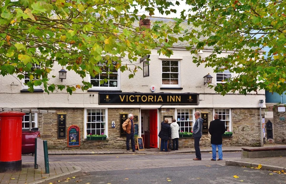 The frontage of the Victoria Inn in Salcombe, a cream and stone pub with a black sign reading Victoria Inn in gold lettering, sash windows, window boxes and people standing outside the red door
