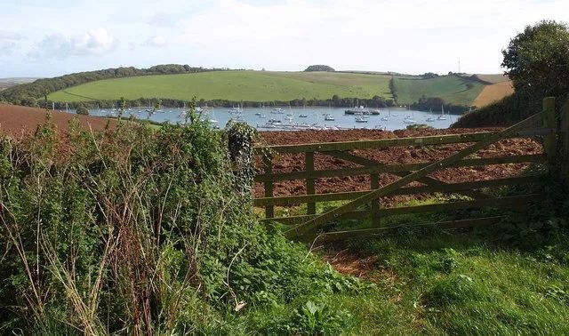 Boats moored on the Kingsbridge Estuary, seen over a farm gate with green fields and wooded hills around the water